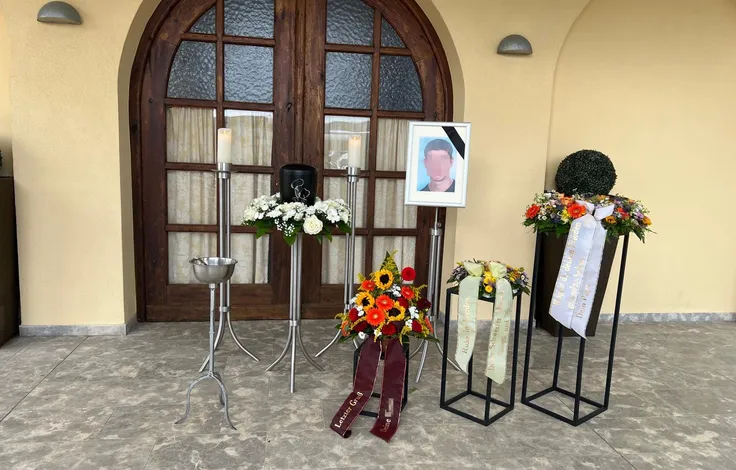 Aufbahrung vor dem Rundbogeneingang einer Trauerhalle: eine dunkle Urne auf einem Ständer zwischen zwei Kerzenleuchtern, daneben das gerahmte Porträt der verstorbenen Person mit Trauerflor, davor Blumengestecke mit Schleifen