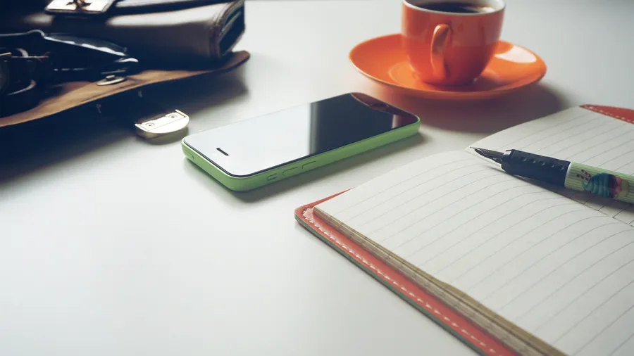 Heller Schreibtisch mit einem aufgeschlagenen Notizbuch samt Stift, einem Smartphone, einer Tasse Kaffee und einer Handtasche am Rand