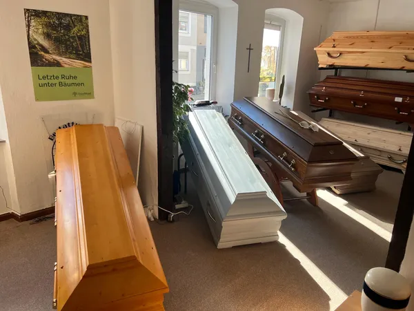 Ausstellungsraum mit mehreren Särgen, an der Wand ein Plakat „Letzte Ruhe unter Bäumen“ und ein schlichtes Holzkreuz