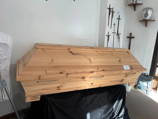 Sarg aus heller Kiefer auf schwarzem Tuch, an der Wand dahinter mehrere Kruzifixe und Grabkreuze
