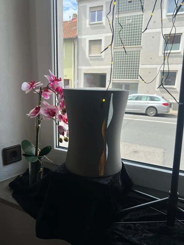 Weiße Urne mit geschwungenem Dekor auf schwarzem Tuch am Fenster, daneben eine Orchidee