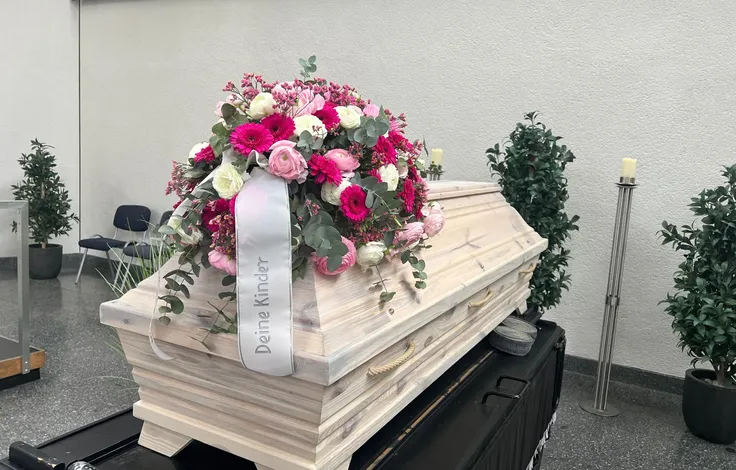 Heller Holzsarg in einer Trauerhalle, darauf ein Gesteck aus pinken Gerbera, Ranunkeln und weißen Rosen mit Eukalyptus