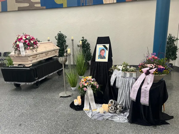 Trauerhalle: heller Sarg mit rosa Blumengesteck, daneben ein Bildrahmen mit Trauerflor, Gestecke und Kerzen
