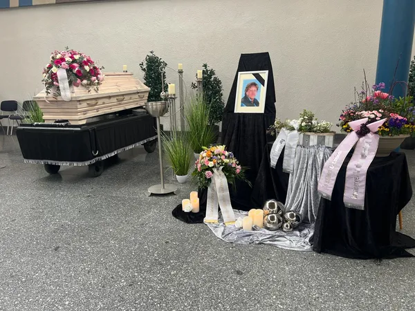 Dieselbe Trauerfeier aus anderem Blickwinkel, mit Sarg, Blumenschmuck und Kerzen auf dunklem Samt