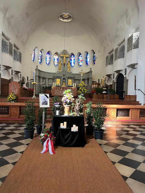 Aufbahrung einer Urne im Mittelgang einer Kirche, mit Blumengesteck, Kerzen und einem Bildrahmen