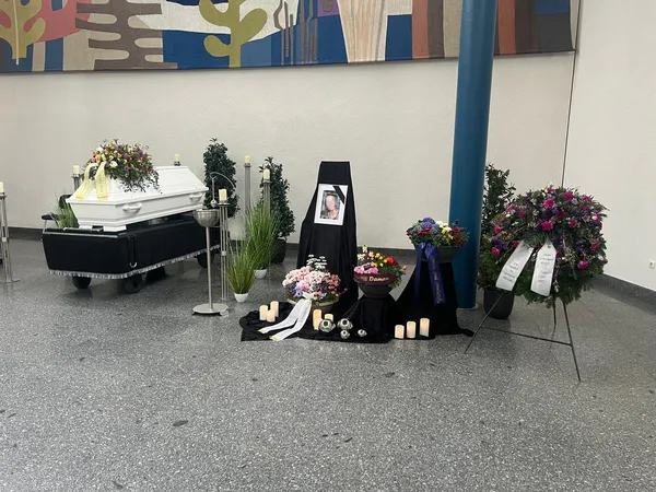Trauerhalle mit weißem Sarg, Blumengestecken und einem Kranz auf einem Ständer